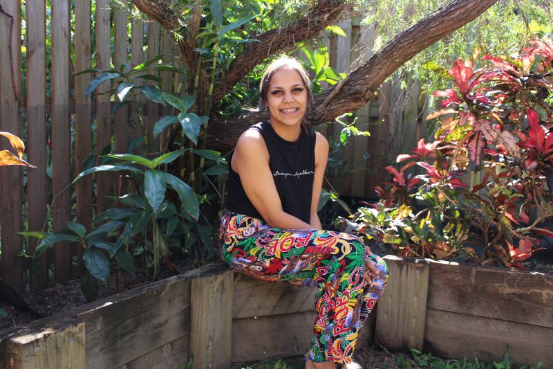 A young women with dark skin sits amongst a garden smiling at the camera.