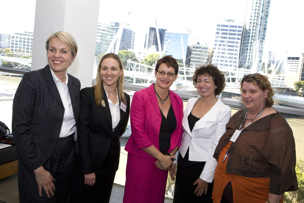 Five women stand around in a group smiling at the camera. They are dressed professionally with the Brisbane skyline behind them.