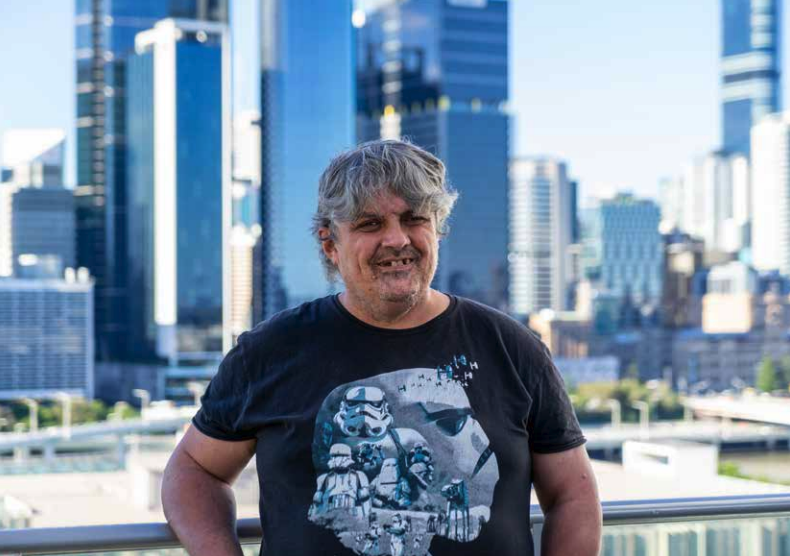 A man smiles at the camera as he stands against the background of the Brisbane City skyline.