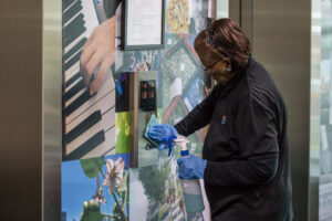 A woman is cleaning the buttons for the elevator using a cleaning cloth. She is wearing a black uniform and blue cleaning gloves. 