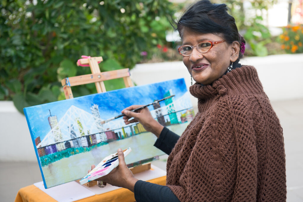 A resident of Brisbane Common Ground stands in the roof top gardens painting a picture. She is turning towards the camera from her painting easel and smiles.