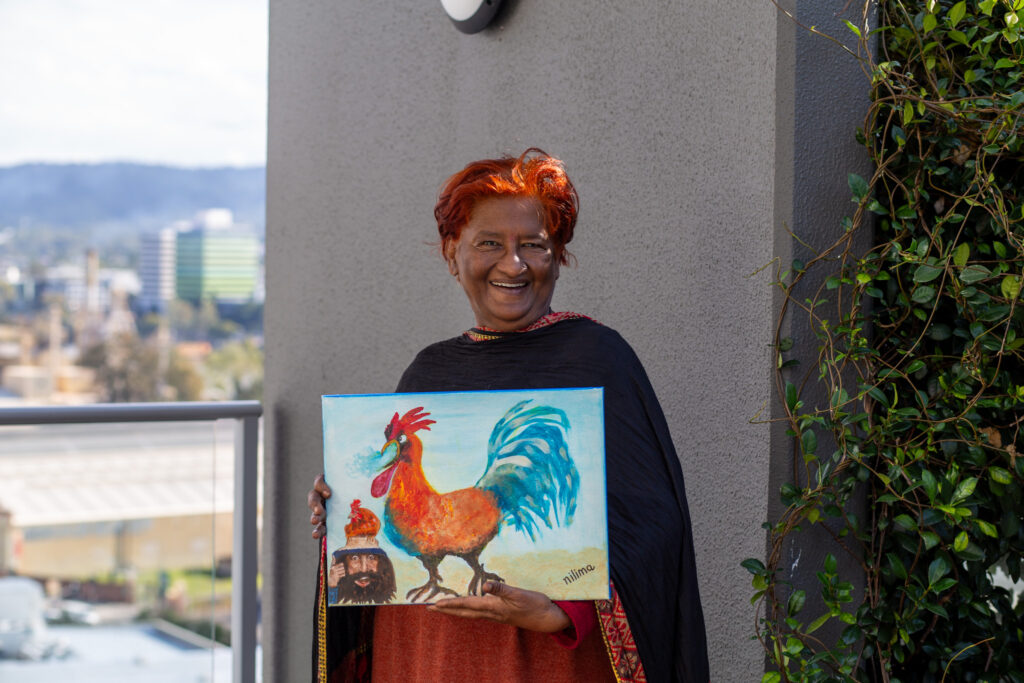 A lady named NIlima holds her painting titled 'Hey CG: Hands off my gal!' The painting is of a rooster inspired by Busan, South Korea and a smaller portrait painting of Costa Georgiadis is placed in the bottom left corner of the canvas.
